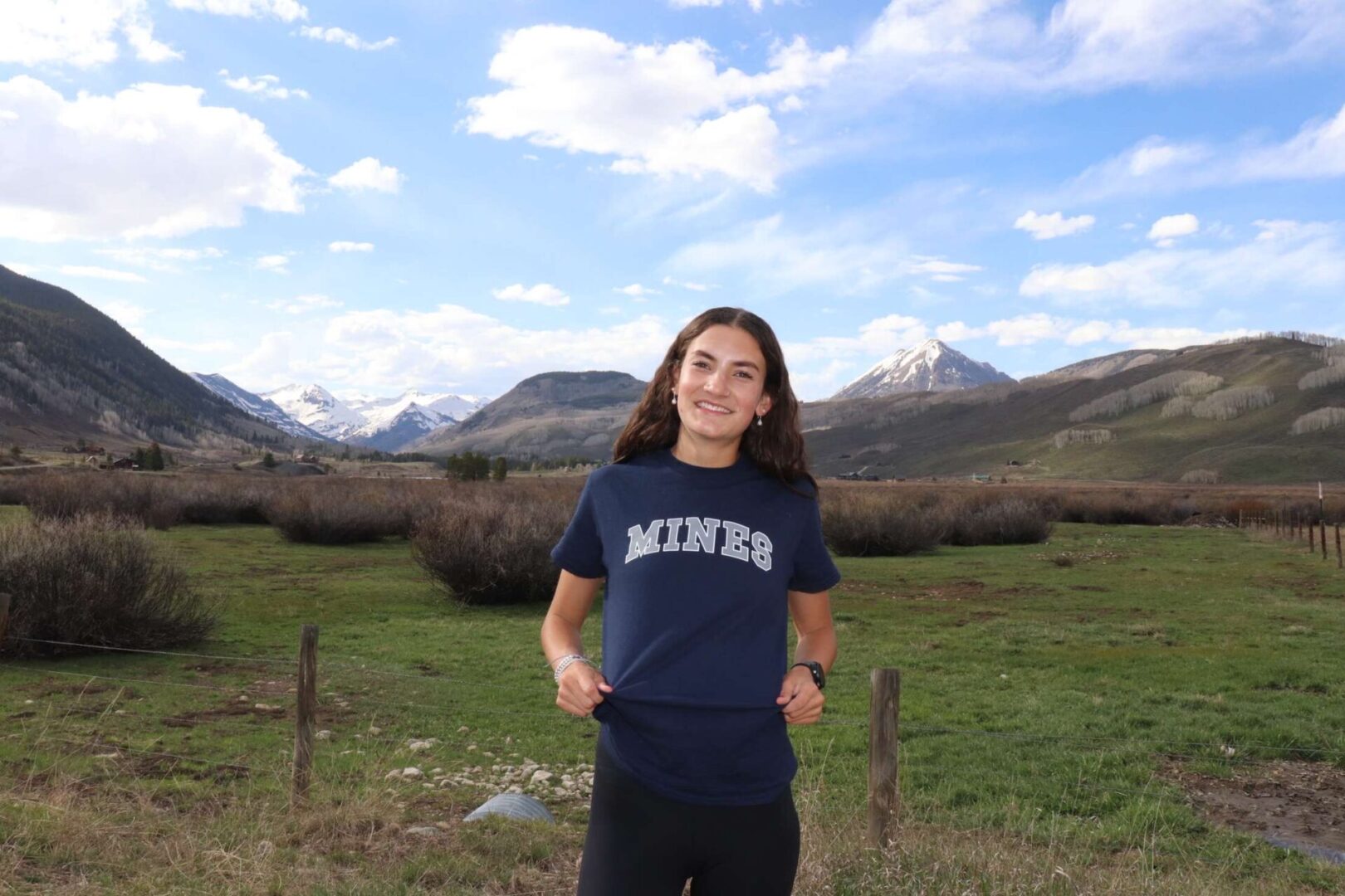 Person smiling in mountainous landscape, wearing MINES shirt.